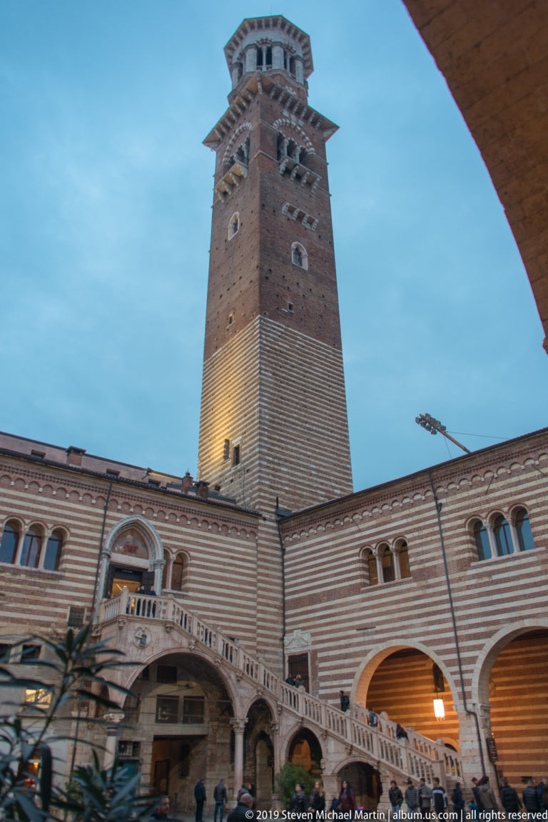 Torre dei Lamberti rising above striped palace arcades at dusk, viewed from the courtyard with people and vehicles below.