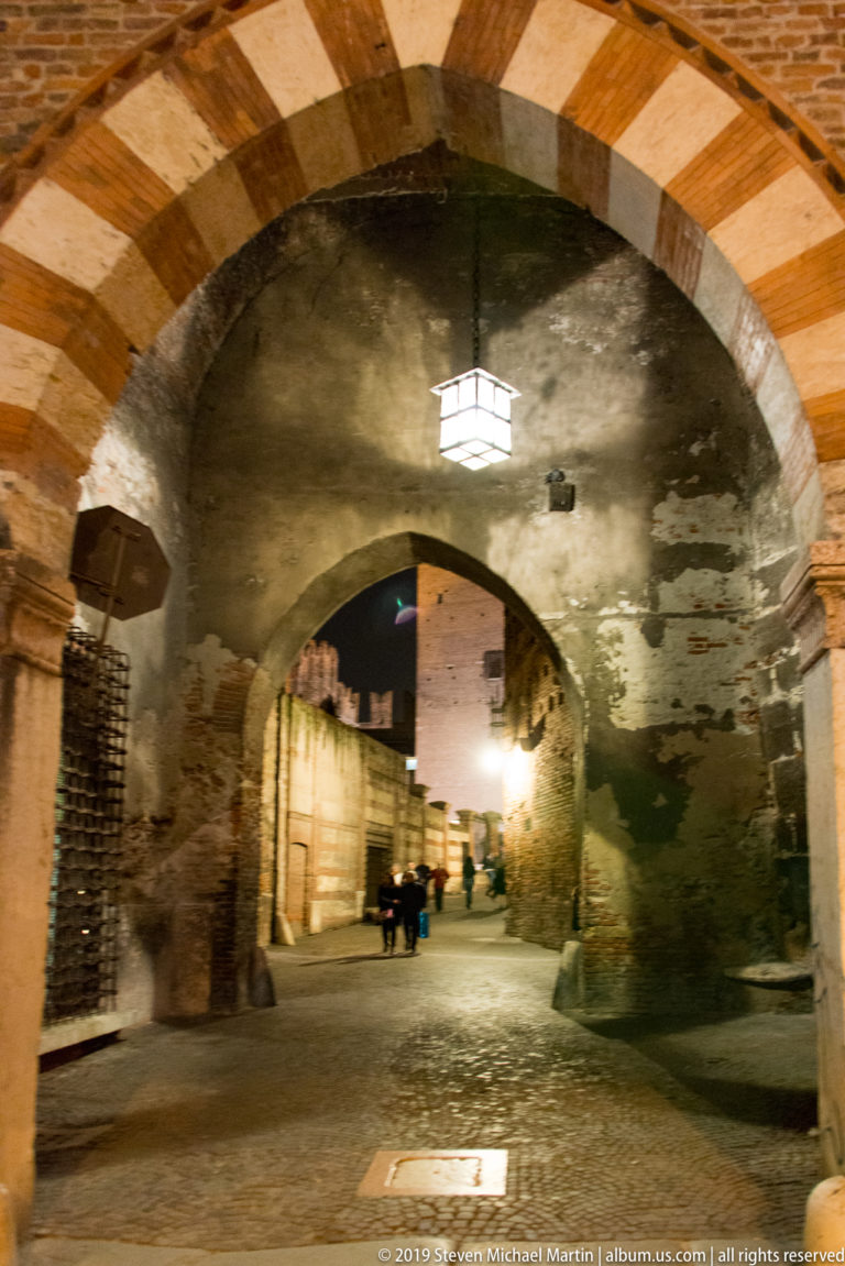 Striped stone arch framing a dim historic passage with rough walls, hanging lantern, iron grilles, and a lit street beyond.