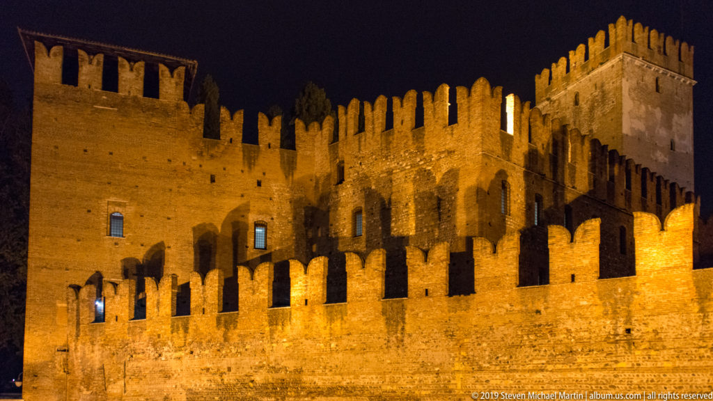 Floodlit brick walls and square tower of Castelvecchio with multiple rows of medieval crenellations against a black sky.