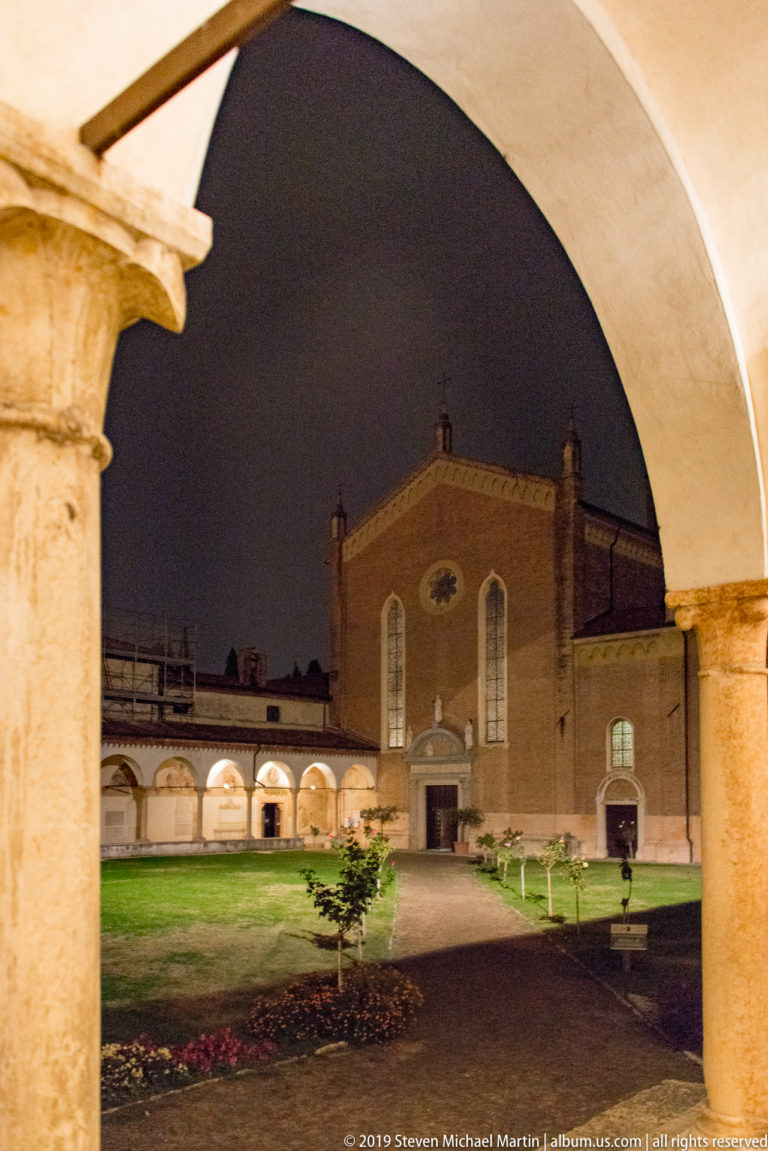 Night view through an arch into a grassy religious courtyard with low arcades and a brick church façade beyond.