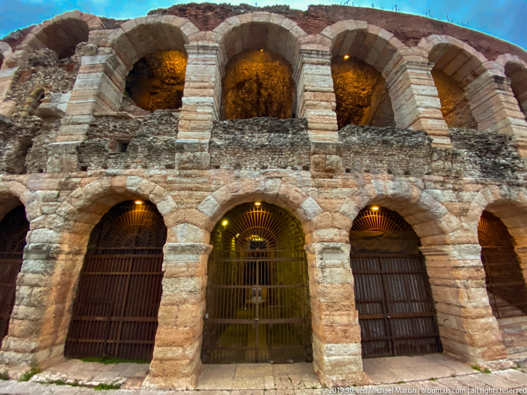 Close view of Verona Arena stone arches with barred entrances below and warmly lit upper openings.