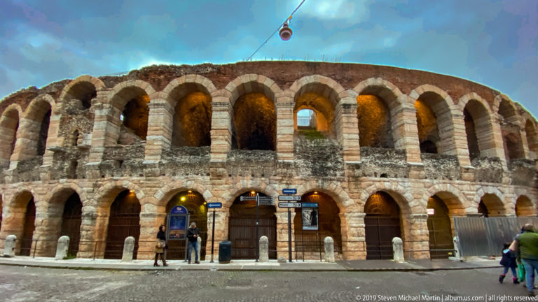Exterior of the Verona Arena with two tiers of weathered stone arches, street signs, bollards, and pedestrians.