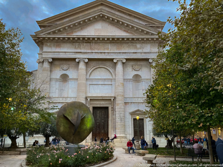 Neoclassical church façade with pediment and columns behind a large dark bronze sphere sculpture in a tree-lined garden.