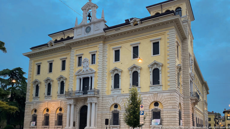 Pale yellow historic civic building with rusticated base, central balcony, pointed window surrounds, rooftop clock and bell, at dusk.