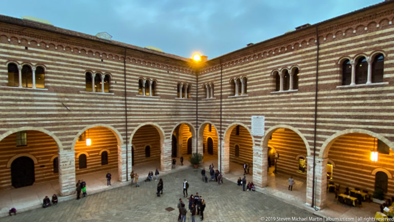 Elevated view into the striped Palazzo della Ragione courtyard with round arcades, upper windows, staircase, lamps, and visitors.