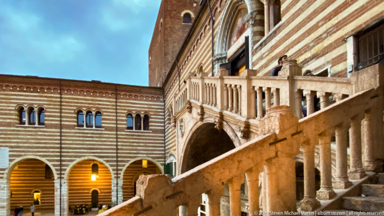 Pale stone Scala della Ragione rising diagonally through a striped brick-and-stone courtyard with arches and upper windows.