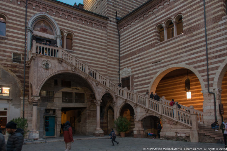 Courtyard view of a broad stone staircase crossing a striped brick palace façade above arcades, with pedestrians below.