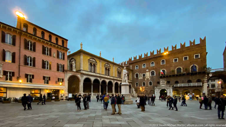 Wide dusk view of Piazza dei Signori with arcaded Renaissance building, crenellated brick palace, stone paving, and pedestrians.