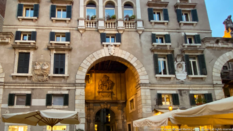 Large illuminated arched passage in an ornate historic façade with shuttered windows, reliefs, balcony, and café umbrellas.