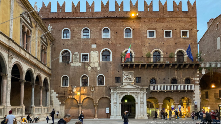 Brick crenellated civic palace with arched windows, white stone portal, balcony flags, arcades, and pedestrians at dusk.