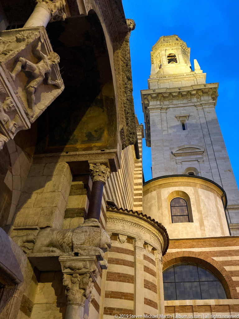 Low-angle view of Verona Cathedral showing striped masonry, a rounded apse, carved stonework, and illuminated bell tower.