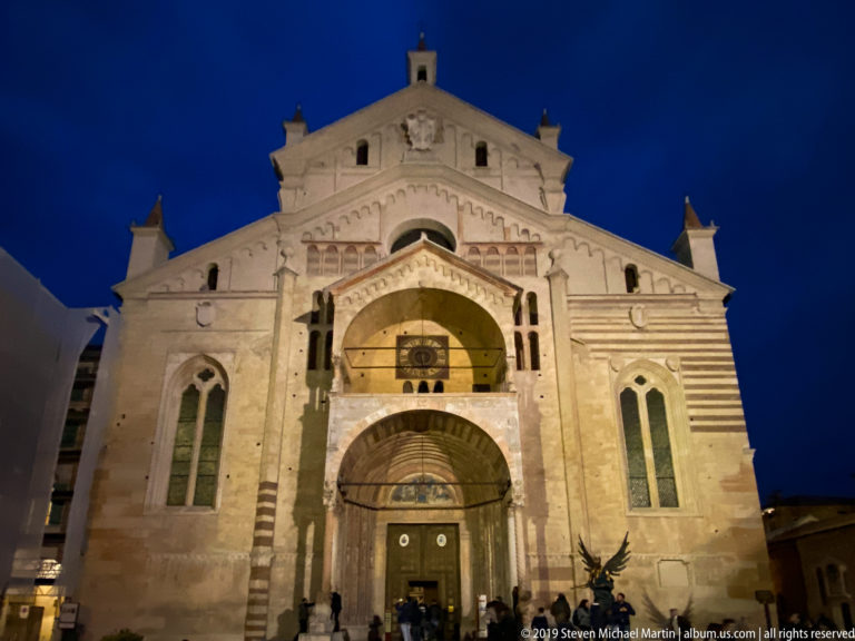 Illuminated pale stone façade of Verona Cathedral with a projecting two-level porch, arched opening, and people at the entrance.