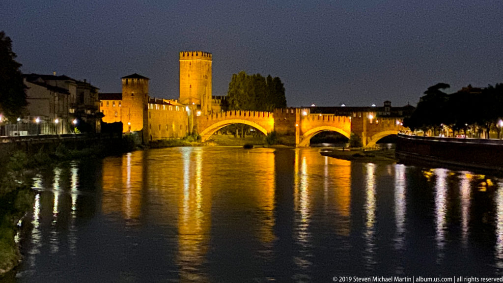 Illuminated Castelvecchio towers and crenellated Ponte Scaligero spanning the Adige, with golden reflections on the river.
