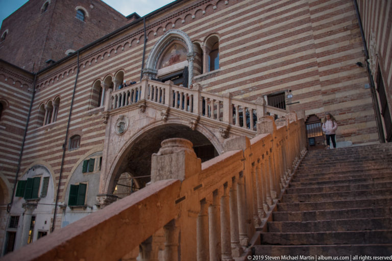 Low-angle view up the monumental exterior staircase against a striped brick-and-stone palace façade, with visitors on the upper steps.