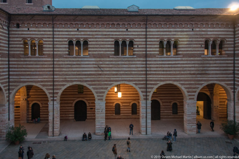 Striped brick-and-stone courtyard with broad arcades, paired upper windows, warm lamps, and scattered visitors.