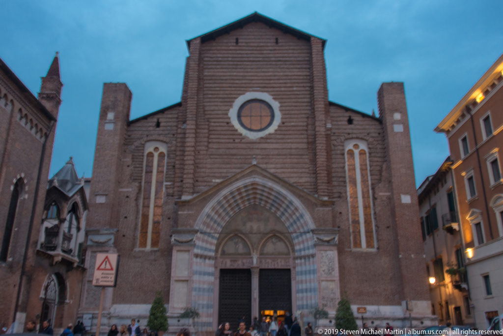 Brick Gothic church façade at dusk with striped pointed portal, circular oculus, tall windows, and people gathered near the entrance.
