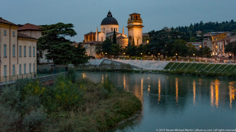Illuminated San Giorgio in Braida dome and bell tower across the Adige, with trees, embankment lights, and reflections.