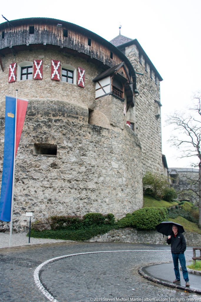 Curved stone wall of Vaduz Castle beside cobbled road, with timber upper story and a person for scale.