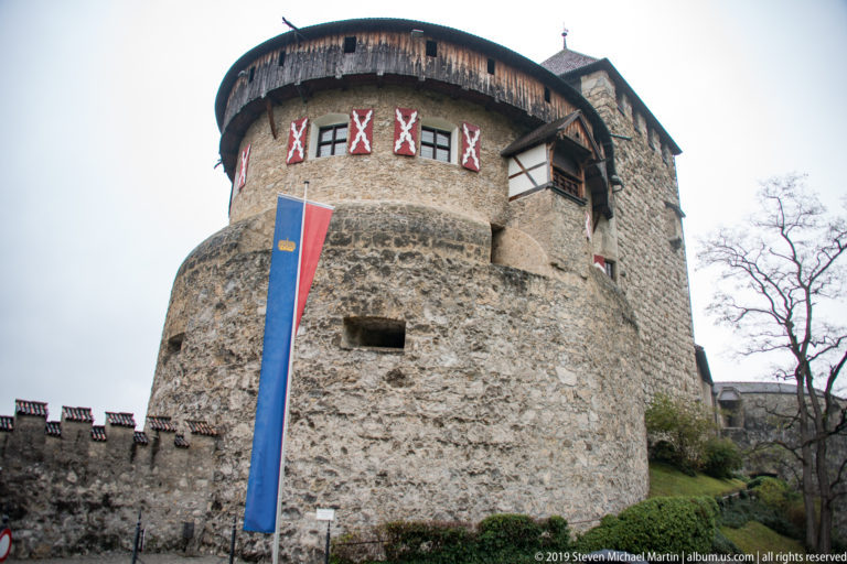 Massive round stone castle tower with timber gallery, patterned shutters, and vertical blue-red banner.