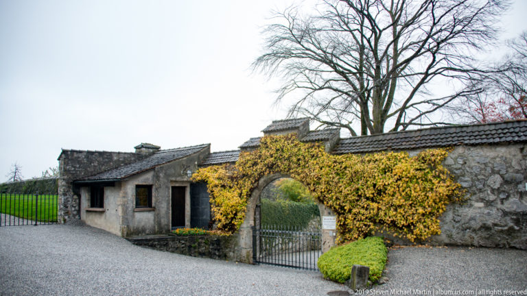 Gravel forecourt with low stone building, ivy-covered arched gate, and large bare tree behind.