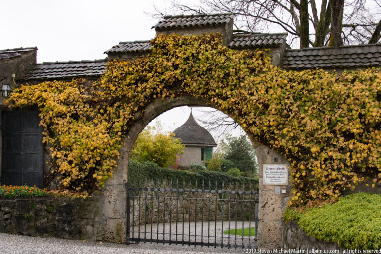 Iron gate beneath an ivy-covered stone arch, with a posted sign and small roofed structure beyond.