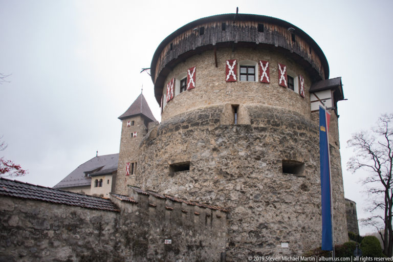 Round tower of Vaduz Castle with patterned shutters and a tall Liechtenstein banner beside the stone wall.