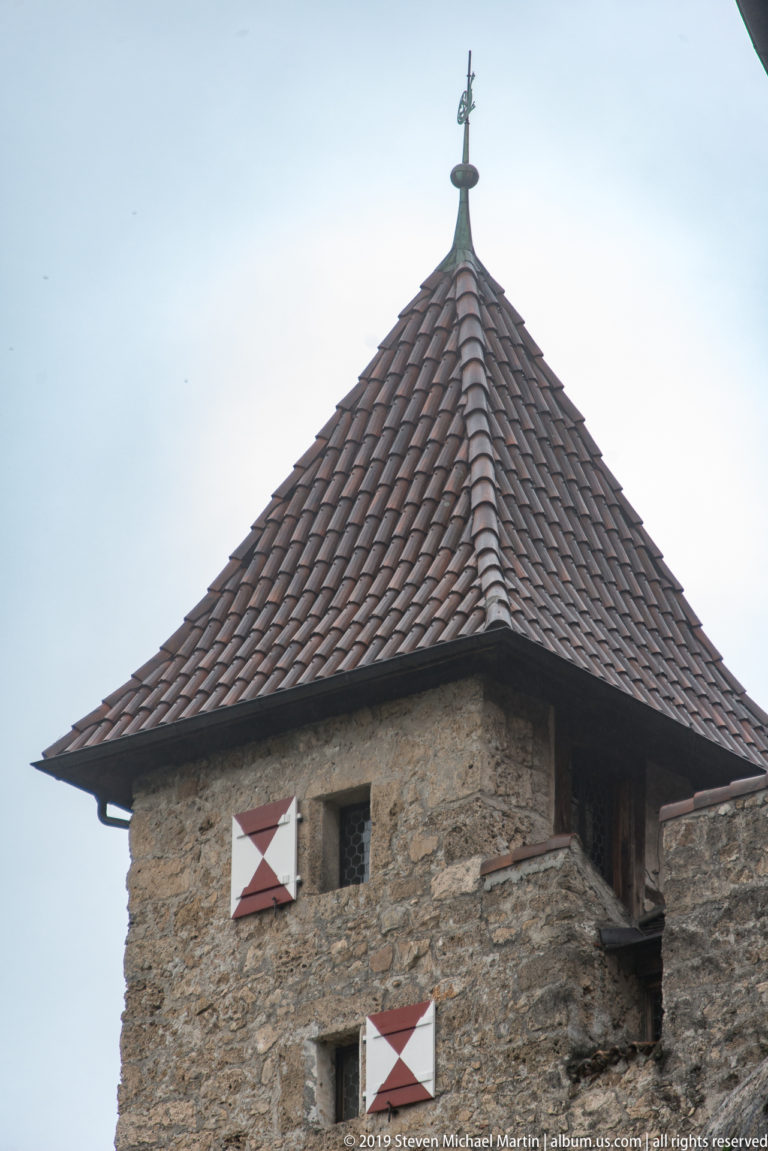 Upper part of a square stone castle tower with hipped tiled roof and red-and-white shutters.