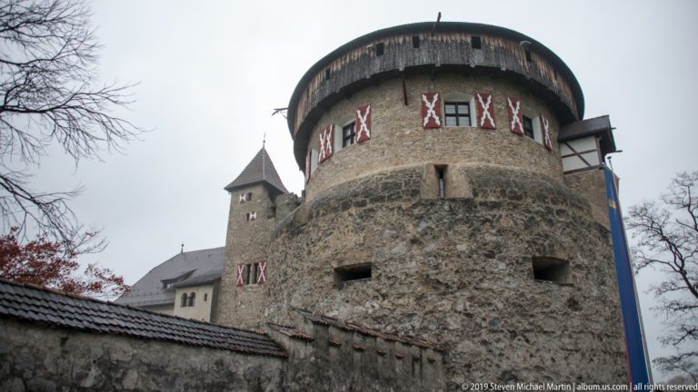Round stone tower of Vaduz Castle behind a perimeter wall, with timber gallery and patterned shutters.
