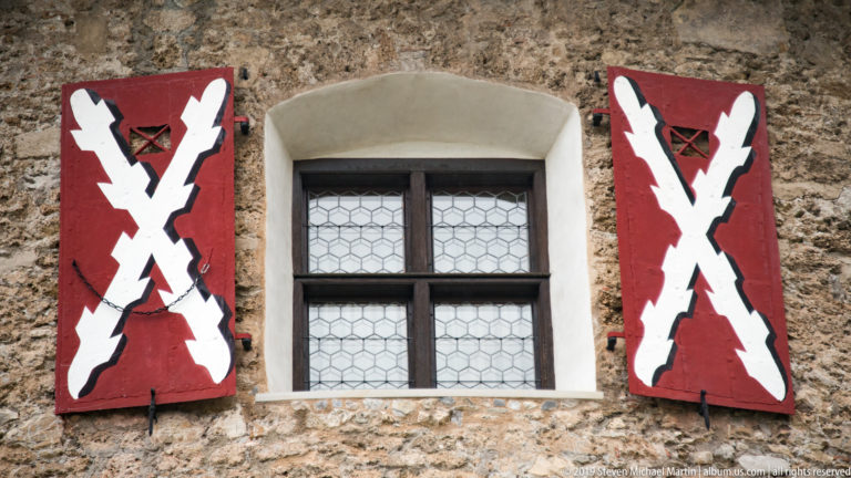Leaded window in rough stone wall flanked by red shutters painted with large white crossed forms.