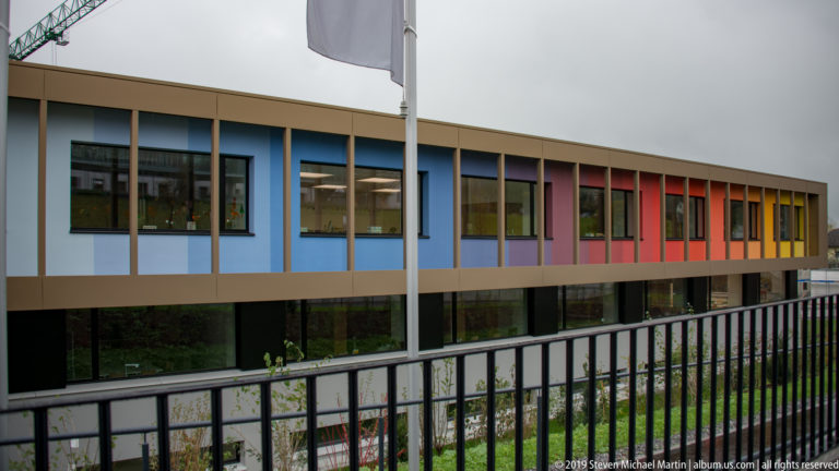 Modern Vaduz building with tall windows and blue, violet, red, orange, and yellow façade panels behind a railing.