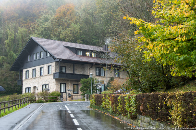 Large pitched-roof hillside house with balconies beside a wet road and autumn trees.