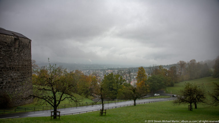 Wet curving road through green hillside with orchard trees and misty Vaduz in the valley beyond.