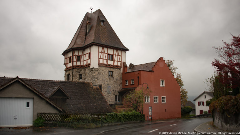 Stone tower with timber-framed upper story and steep roof beside an ochre-red house on a quiet street.