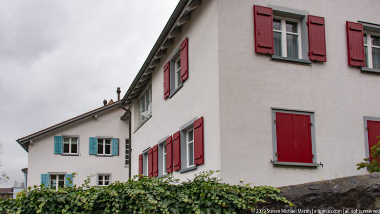 White houses with red and blue-green shutters, steep roofs, and ivy along the foreground wall.