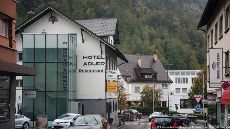Herrengasse street with Hotel Adler sign, cars, pedestrians, shops, and wooded hillside beyond.