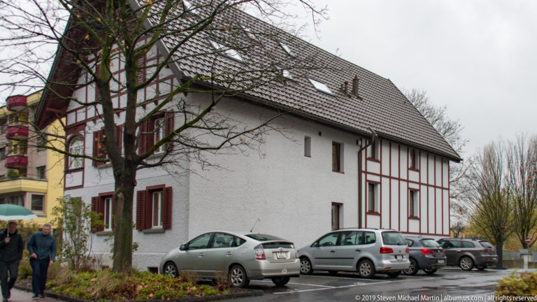 White steep-roofed building with red-brown timber framing and parked cars along one side.