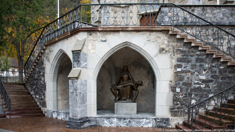 Dark figurative sculpture in a pointed-arch niche beneath stone stairs and curved iron railings.