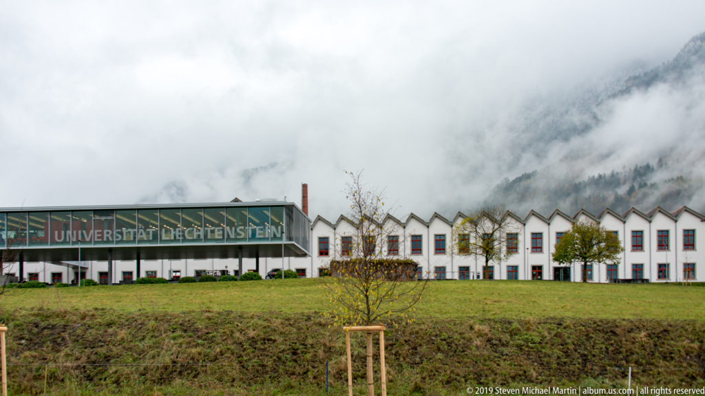 University of Liechtenstein buildings with a long glazed façade and repeated white gables beneath misty mountain slopes.