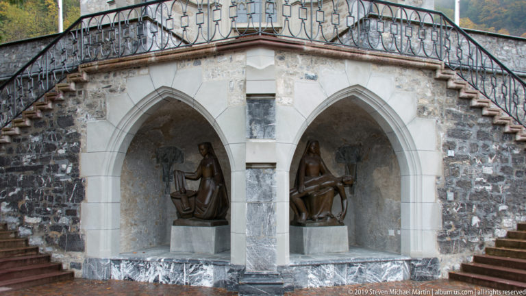 Two dark figurative sculptures in adjacent pointed-arch stone niches beneath an exterior railing.