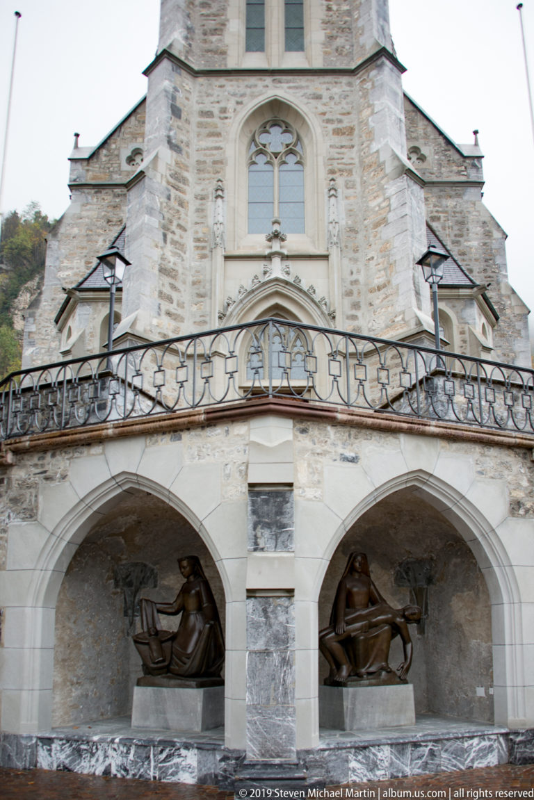 Two pointed-arch niches with dark sculptures beneath stairs on the stone façade of St. Florin Cathedral.