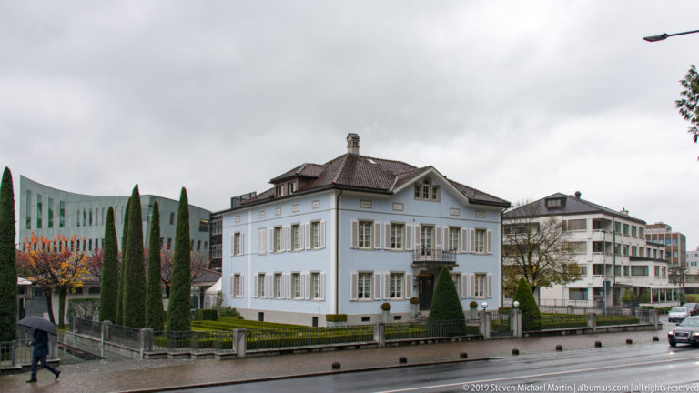 White hipped-roof villa with green shutters and formal garden beside a rainy street in Vaduz.