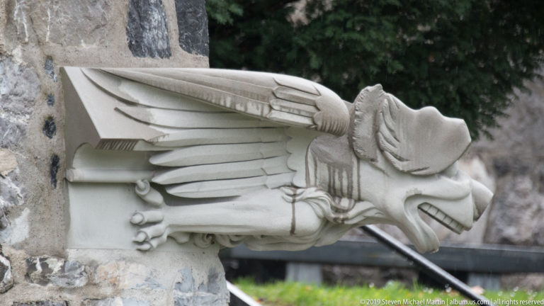 Side view of a pale carved winged human figure projecting horizontally from a stone wall.