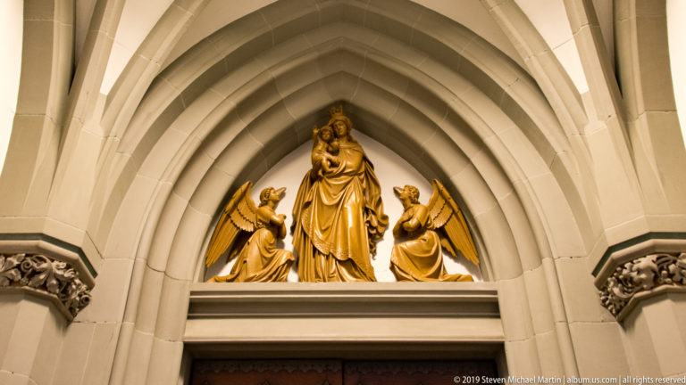 Gilded crowned Virgin holding the Christ Child between two kneeling angels in a pointed stone doorway tympanum.