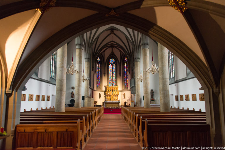 St. Florin Cathedral nave framed by a dark pointed arch, with pews, columns, chandeliers, stained glass, and altar beyond.