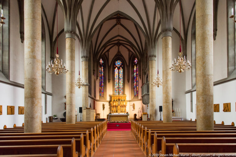 Central nave of St. Florin Cathedral with wooden pews, stone columns, chandeliers, ribbed vaults, and high altar ahead.