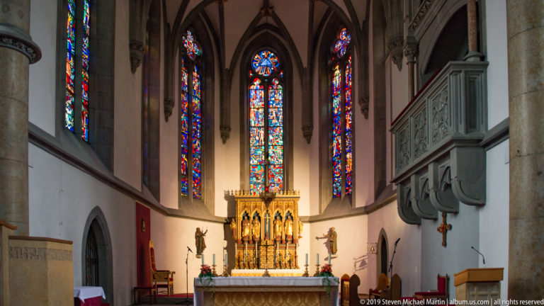 Gilded high altar beneath three tall stained-glass windows, with statues and carved pulpit in St. Florin Cathedral.