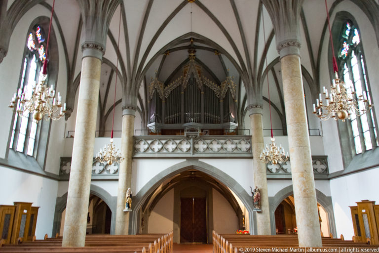 Rear view of St. Florin Cathedral nave with tall columns, organ gallery, chandeliers, stained glass, and central entrance arch.