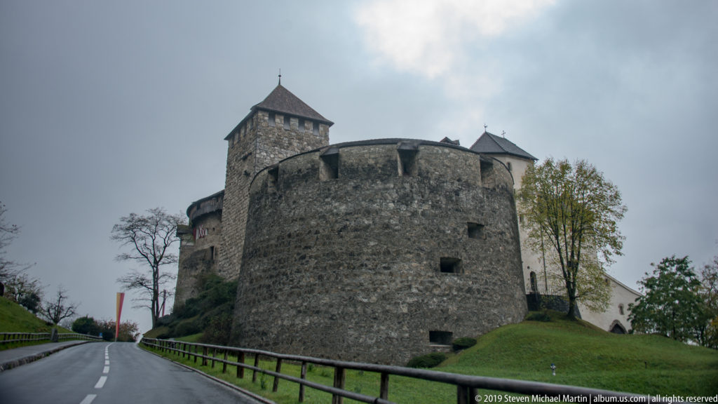 Stone walls and towers of Vaduz Castle rise beside a curving uphill road under an overcast sky.