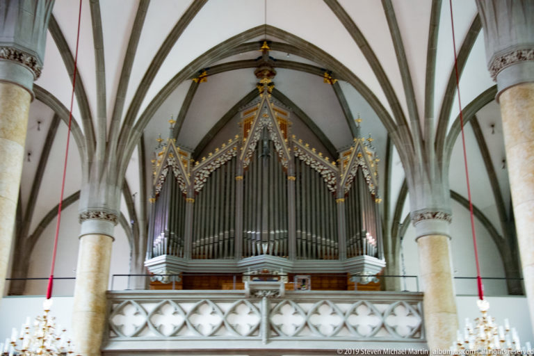 Frontal view of the St. Florin Cathedral pipe organ between stone columns, ribbed vaults, and a pierced gallery rail.