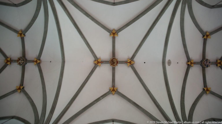 Pale church ceiling crossed by gray ribs converging at small gilded and painted bosses.